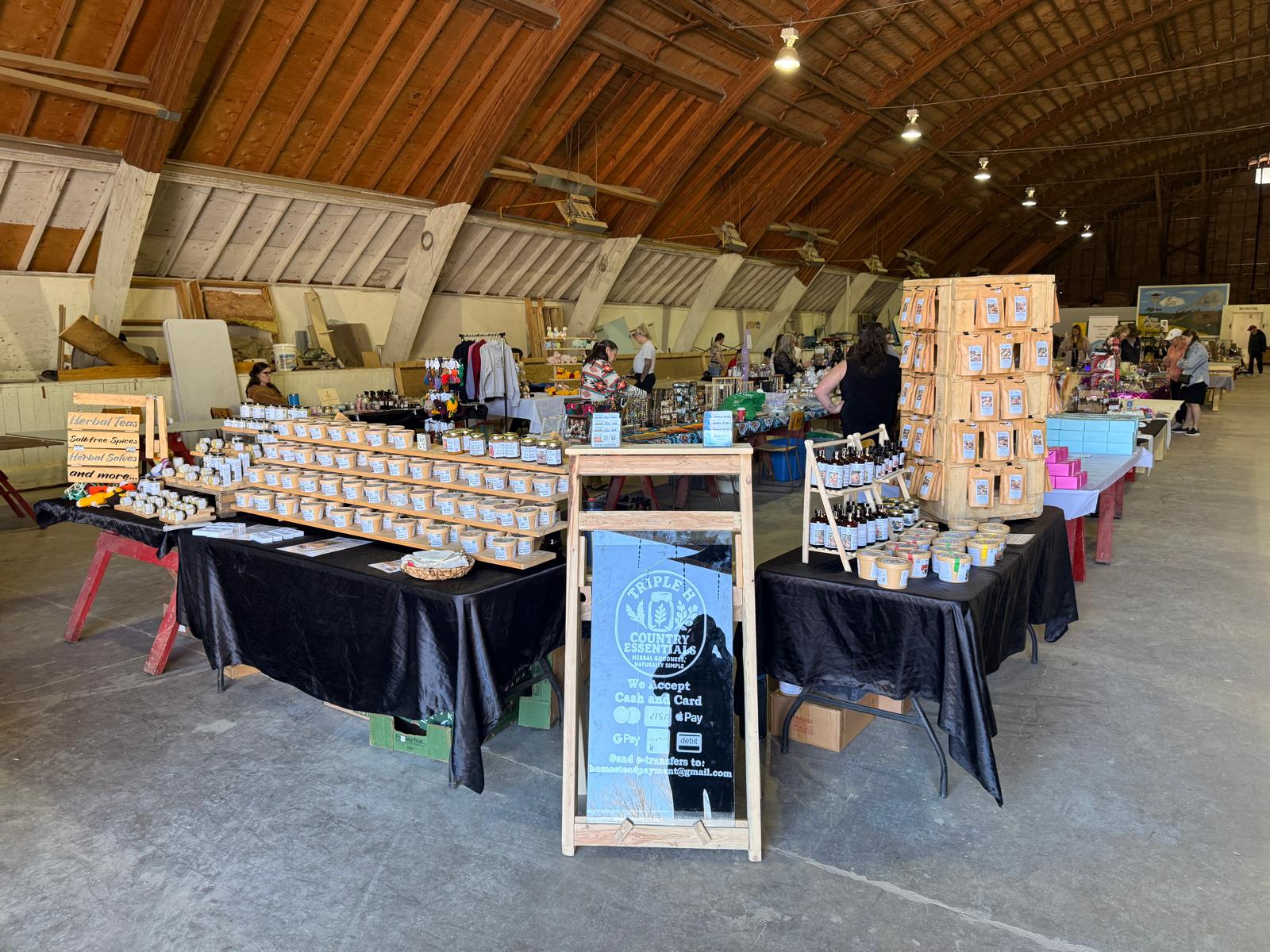 Indoor market or fair with tables displaying goods and a sign in the center.