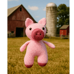 Crocheted pink pig toy in a field with a barn and silo in the background